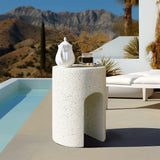 An ivory white cement side table, boasting a sleek U-shaped design, holds a teapot, coffee cup, and decorative branches beside a pool with mountains in the background.
