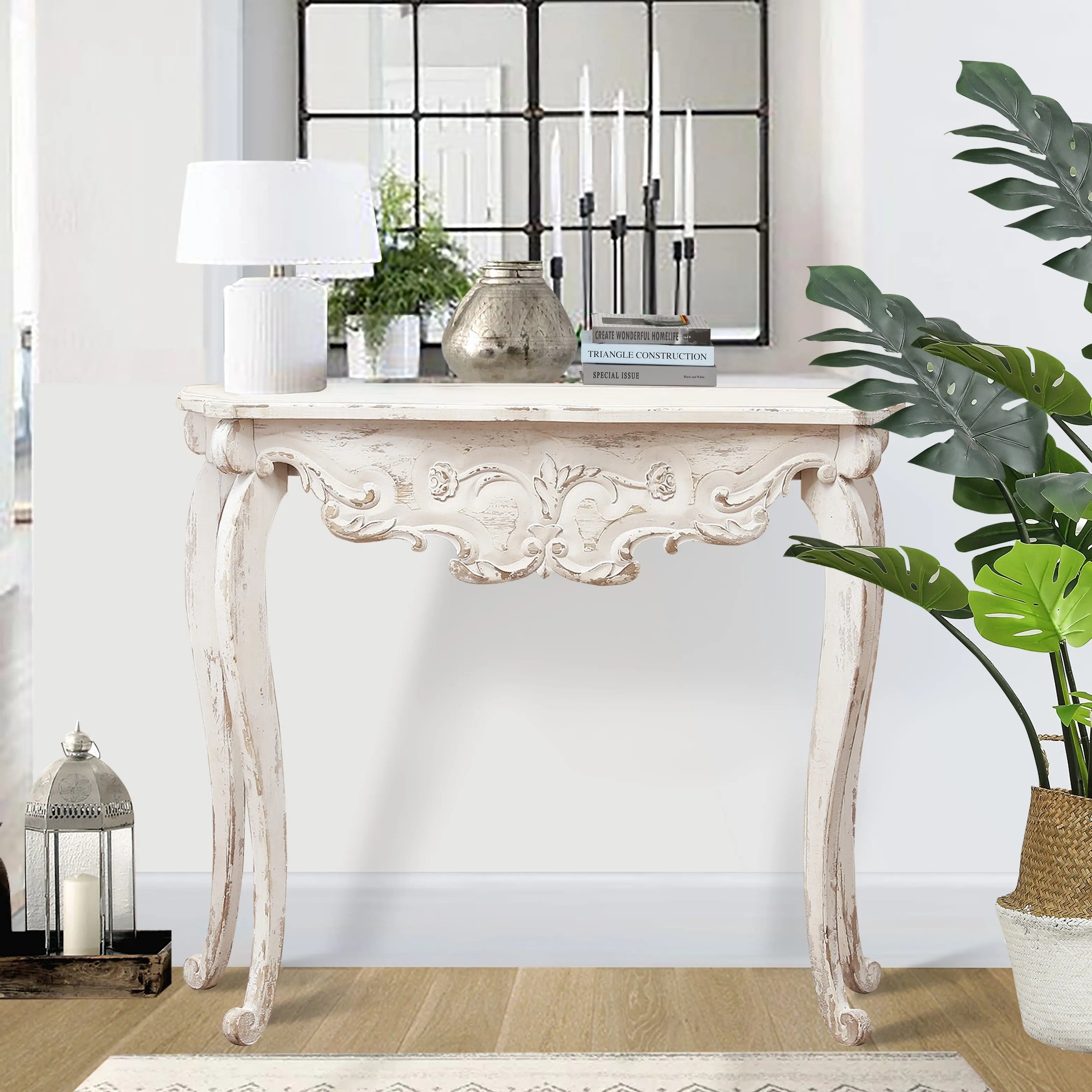 A Victorian-style console table with a distressed off white finish features ornate carvings and holds books, a lamp, a candle holder, and decor, all arranged in front of a mirror with plants nearby.