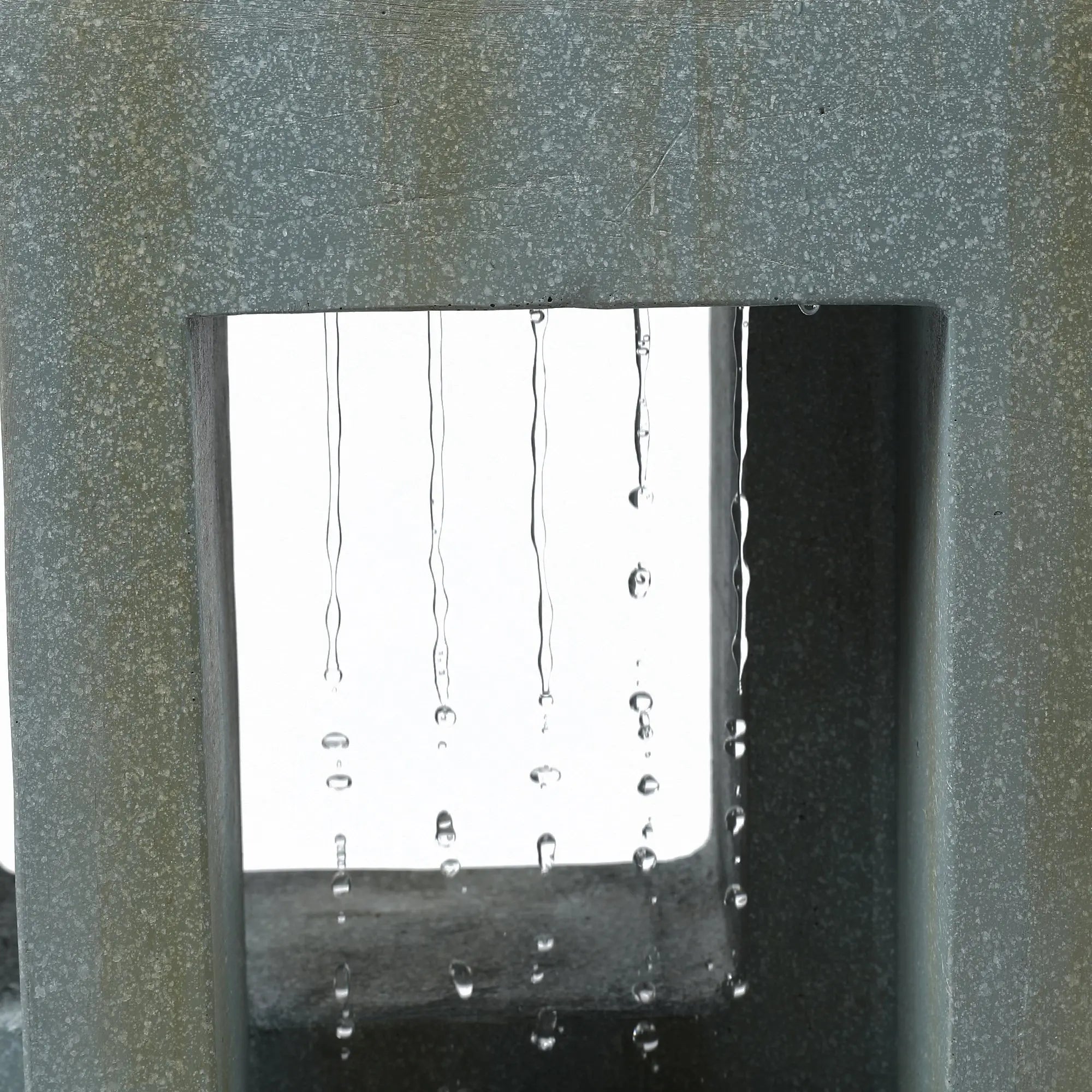 Close-up of water droplets falling in straight lines through a rectangular opening in a textured grey structure, creating a striking water fountain feature perfect for any outdoor living space.