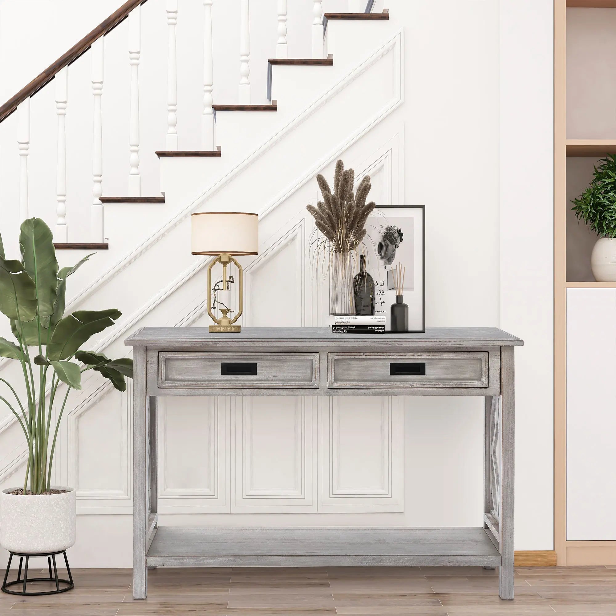 A distressed gray washed console table with two drawers stands under a staircase, decorated with a lamp, framed art, vases, and dried plants. A potted plant sits to the left of this chic piece.