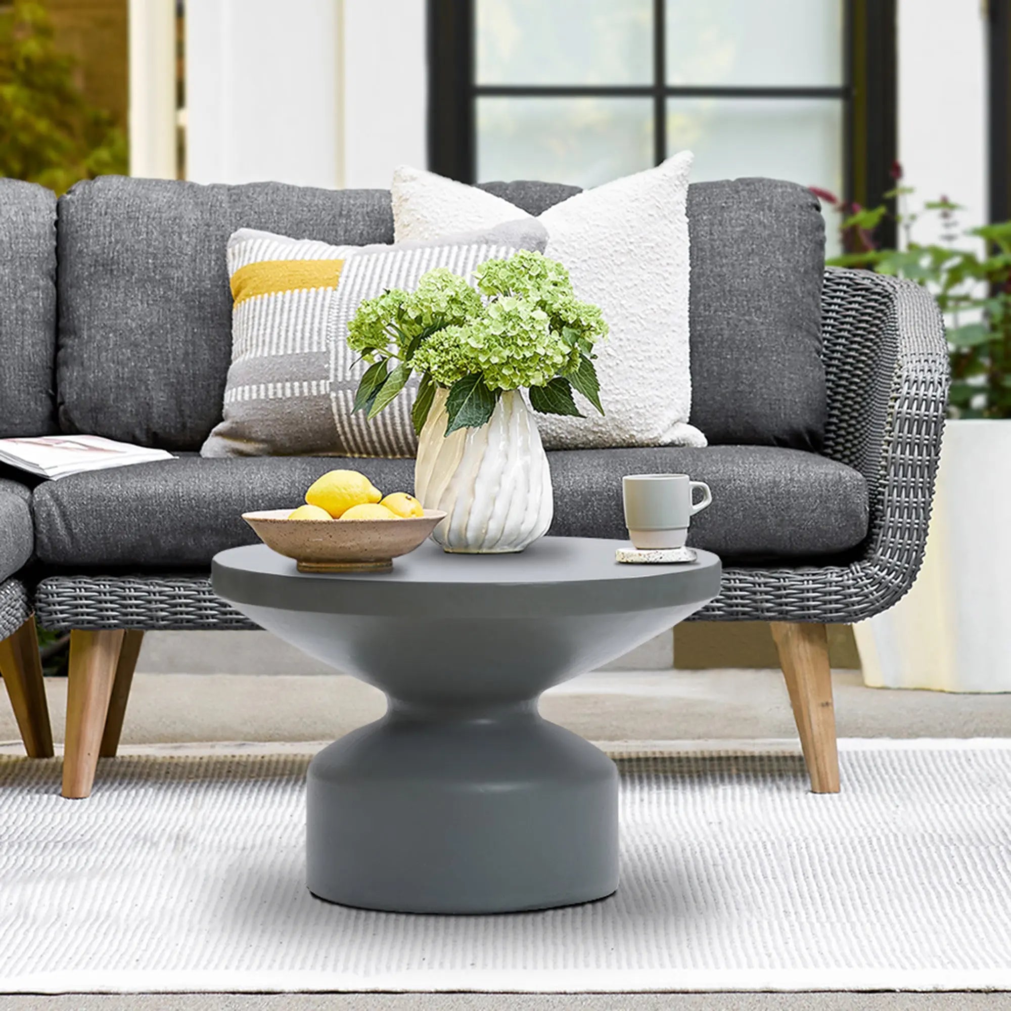 A contemporary round coffee table with a gray finish, crafted from magnesium oxide material, displays green hydrangeas, lemons, and a gray mug in front of a woven sofa with patterned cushions.