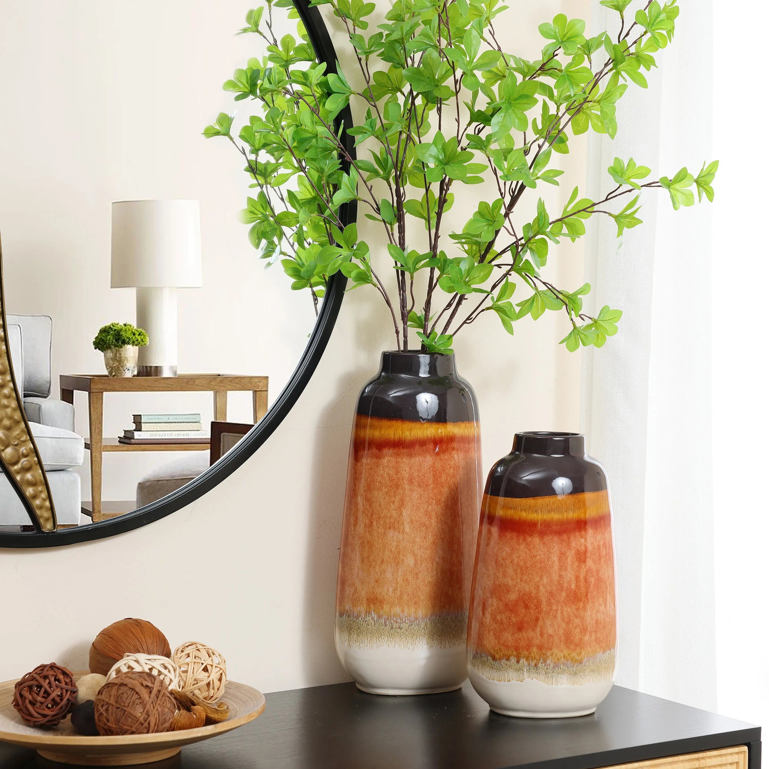 Two vintage ceramic vases with orange tones, one taller with green branches, sit elegantly on a dark table. A round mirror and lamp on a wooden table add charm to the home decor, reflecting beautifully in the background.