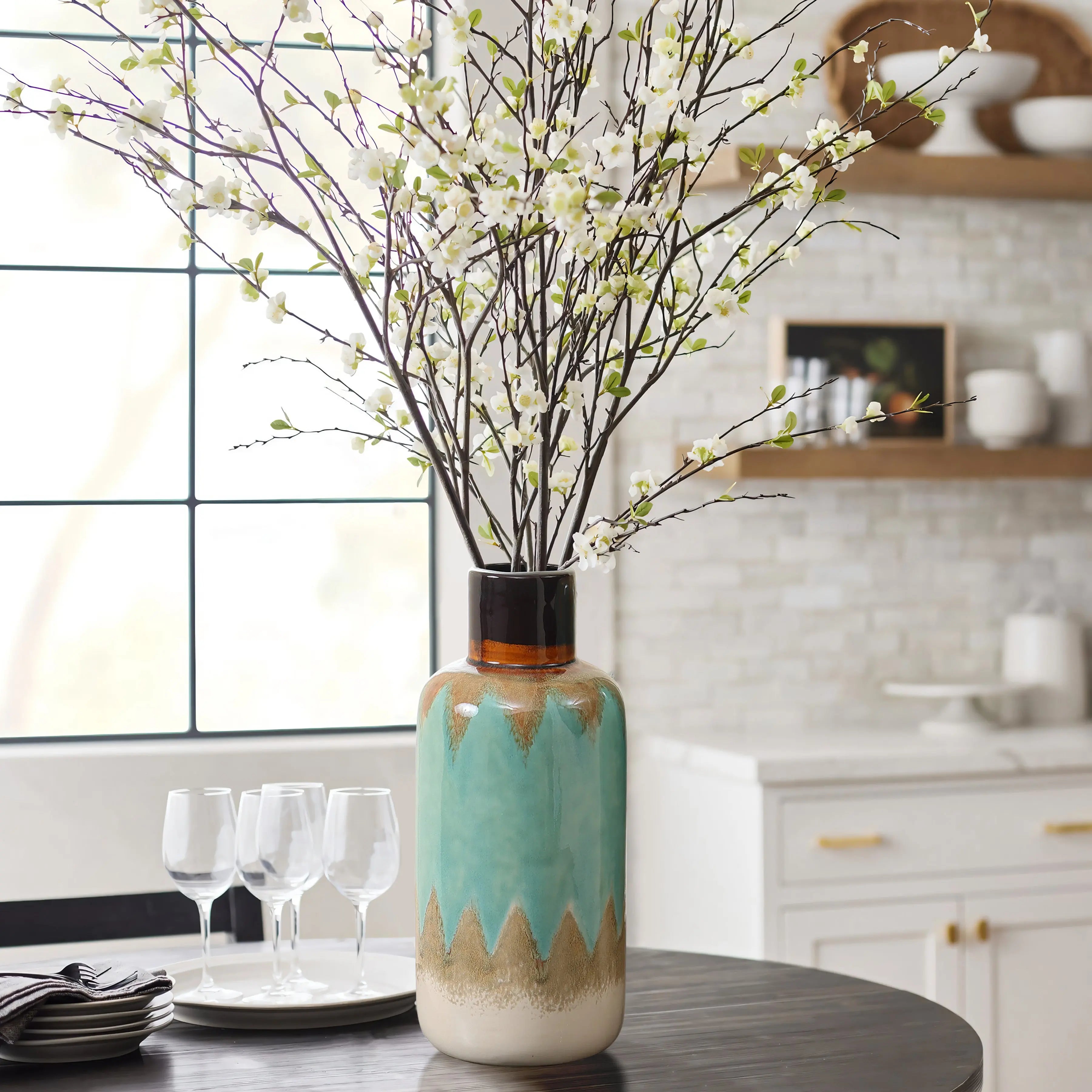A coastal-inspired vase with a zigzag pattern holds white flowering branches on a table set with four wine glasses in a kitchen.
