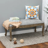 A wooden upholstered bench with linen upholstery, a gray cushion, beige throw, books, and a floral pillow sits on a patterned rug; woven balls rest nearby on the floor, adding old-world charm to the space.