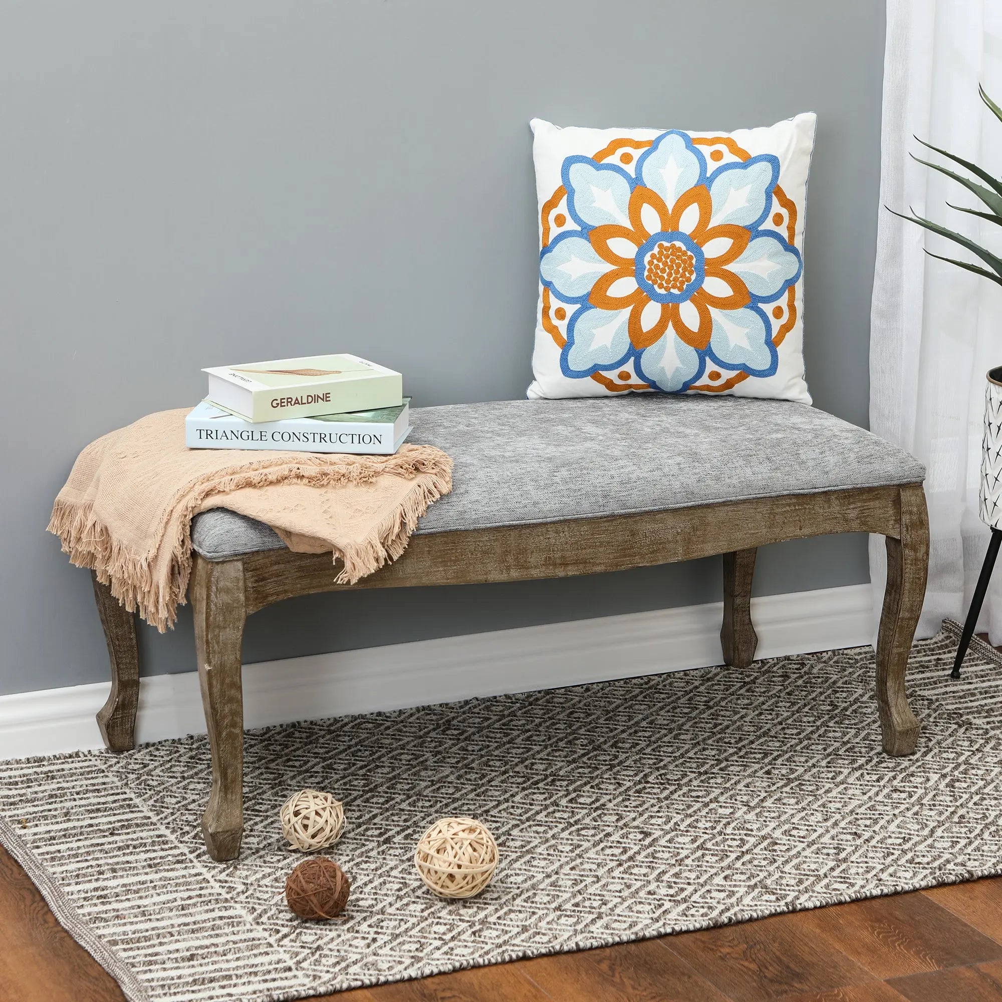 A wooden upholstered bench with linen upholstery, a gray cushion, beige throw, books, and a floral pillow sits on a patterned rug; woven balls rest nearby on the floor, adding old-world charm to the space.