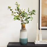 A unique ceramic vase bursting with colorful flowering branches is placed on a dark table next to two unlit white candles.