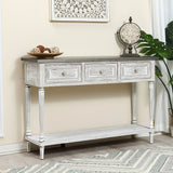 A farmhouse-inspired table, this white distressed wood console features three drawers, a lower shelf, and is adorned with potpourri, an ornate clock, and a large green plant nearby.