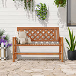 A solid wood garden bench adorned with decorative cushions graces a stone patio, surrounded by potted plants and hanging planters against a crisp white wall.