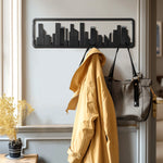 A coat and bag hang on a wall-mounted rack crafted from solid fir wood, its sleek design mimicking a city skyline. Flip-down hooks add functionality, while a nearby potted plant brings a touch of nature to the urban-inspired decor.