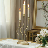 A three-armed gold candle holder with lit candles graces a table adorned with a white cloth and a vase of white flowers, set against the elegant home decor backdrop of a framed painting and curtains.