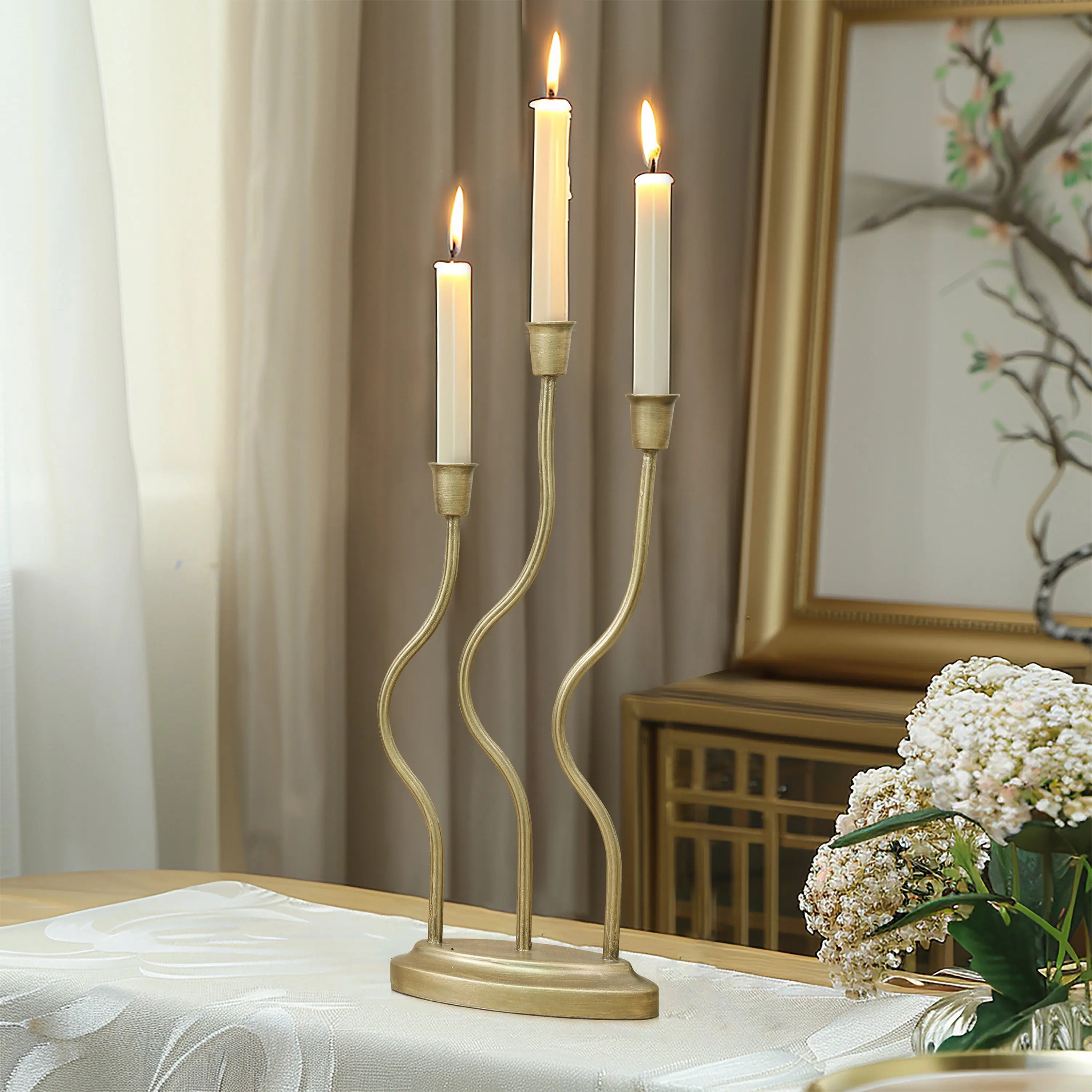 A three-armed gold candle holder with lit candles graces a table adorned with a white cloth and a vase of white flowers, set against the elegant home decor backdrop of a framed painting and curtains.
