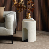 A textured ivory white cement side table, with a sleek U-shaped design, holds a vase with dried leaves and a stack of books next to a light gray sofa in a room adorned with wooden paneling and a beige patterned rug.
