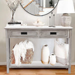 A distressed gray washed console table with two drawers, decorated with a lamp, vases, candles, books, a bowl, and a woven basket with a blanket and pillow, stands against a wall topped by a round mirror.