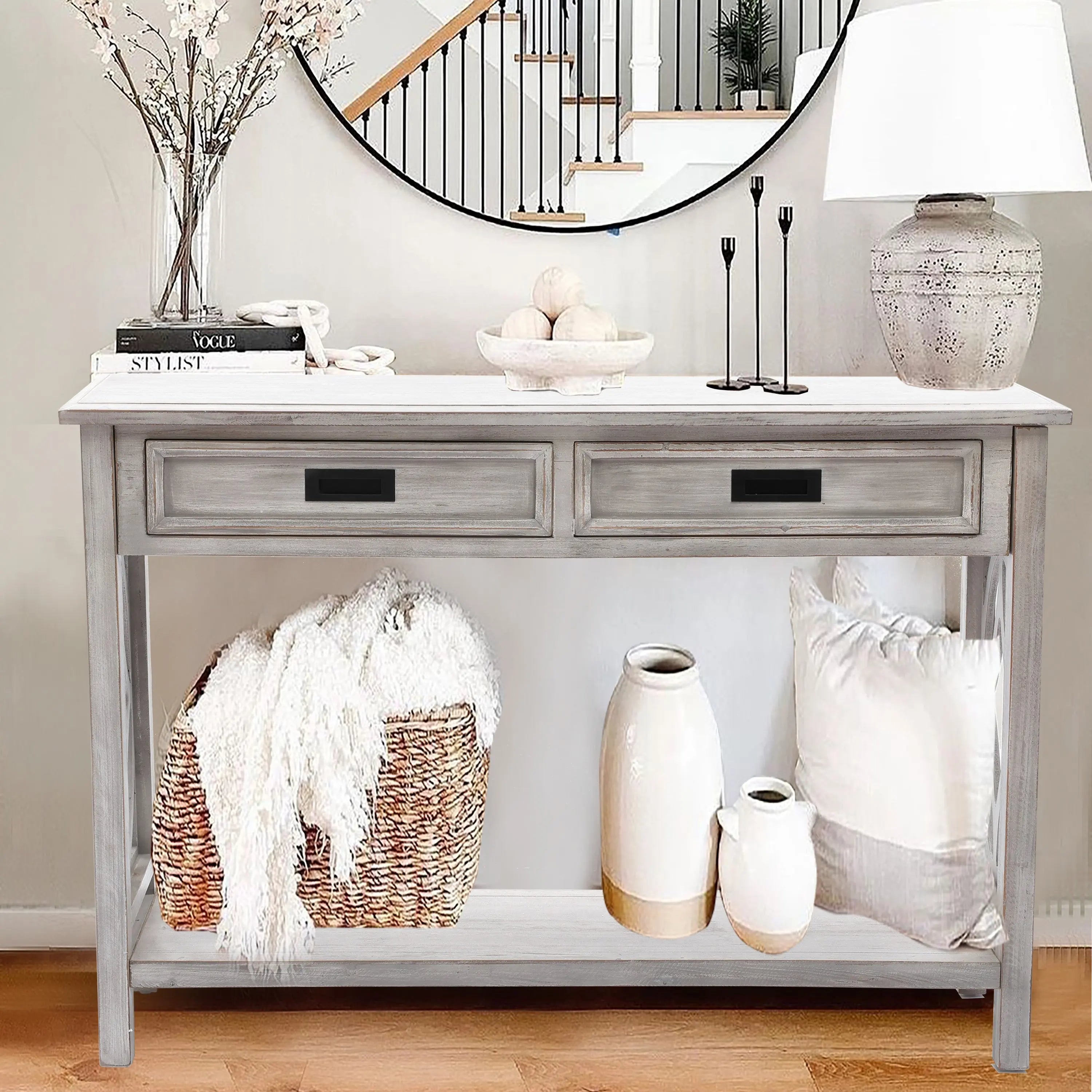 A distressed gray washed console table with two drawers, decorated with a lamp, vases, candles, books, a bowl, and a woven basket with a blanket and pillow, stands against a wall topped by a round mirror.