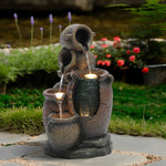 Three stone jugs form a rustic urn outdoor fountain with built-in LED light, set on stone tiles in a garden with grass, flowers, and a pond in the background—an elegant addition to your garden decor.