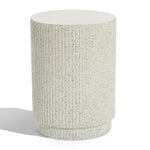 A textured, cylindrical, light-speckled concrete pedestal with vertical ridges stands on a flat surface, casting a shadow. This versatile table effortlessly blends function and style, offering an elegant touch to any space.