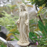 A white Mary statue in a robe stands outdoors among green plants and flowers, her arms gently extended and palms up—a serene religious icon crafted from durable magnesium oxide.