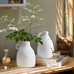 Vintage-inspired design meets modern elegance as white flower vases and a small terracotta vase with tall greenery sit gracefully on the table. Nearby, books and a bowl with a spoon bask in the sunlight streaming through the window.