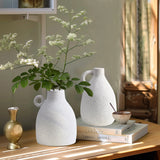 Vintage-inspired design meets modern elegance as white flower vases and a small terracotta vase with tall greenery sit gracefully on the table. Nearby, books and a bowl with a spoon bask in the sunlight streaming through the window.