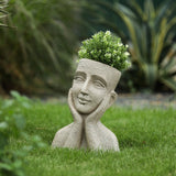 A bust planter shaped like a human head with hands on its cheeks, containing a green leafy plant, sits on grass in a garden, creating the look of a decorative statue.