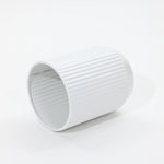 A sleek, white, cylindrical object with vertical ridges lies on its side against a white background, reminiscent of modern metal plant pots in its streamlined design.