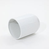A sleek, white, cylindrical object with vertical ridges lies on its side against a white background, reminiscent of modern metal plant pots in its streamlined design.