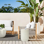 Outdoor seating area with two white cushioned chairs and an ivory white side table, crafted from durable cement material. A large potted plant and trees grace the background under a clear blue sky, offering a serene retreat.