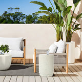 Outdoor seating area with two white cushioned chairs and an ivory white side table, crafted from durable cement material. A large potted plant and trees grace the background under a clear blue sky, offering a serene retreat.